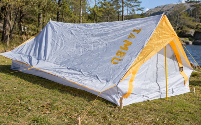 Tenda grigia con accenti gialli e scritta gialla 'KAMBU' su erba vicino a un lago e alberi