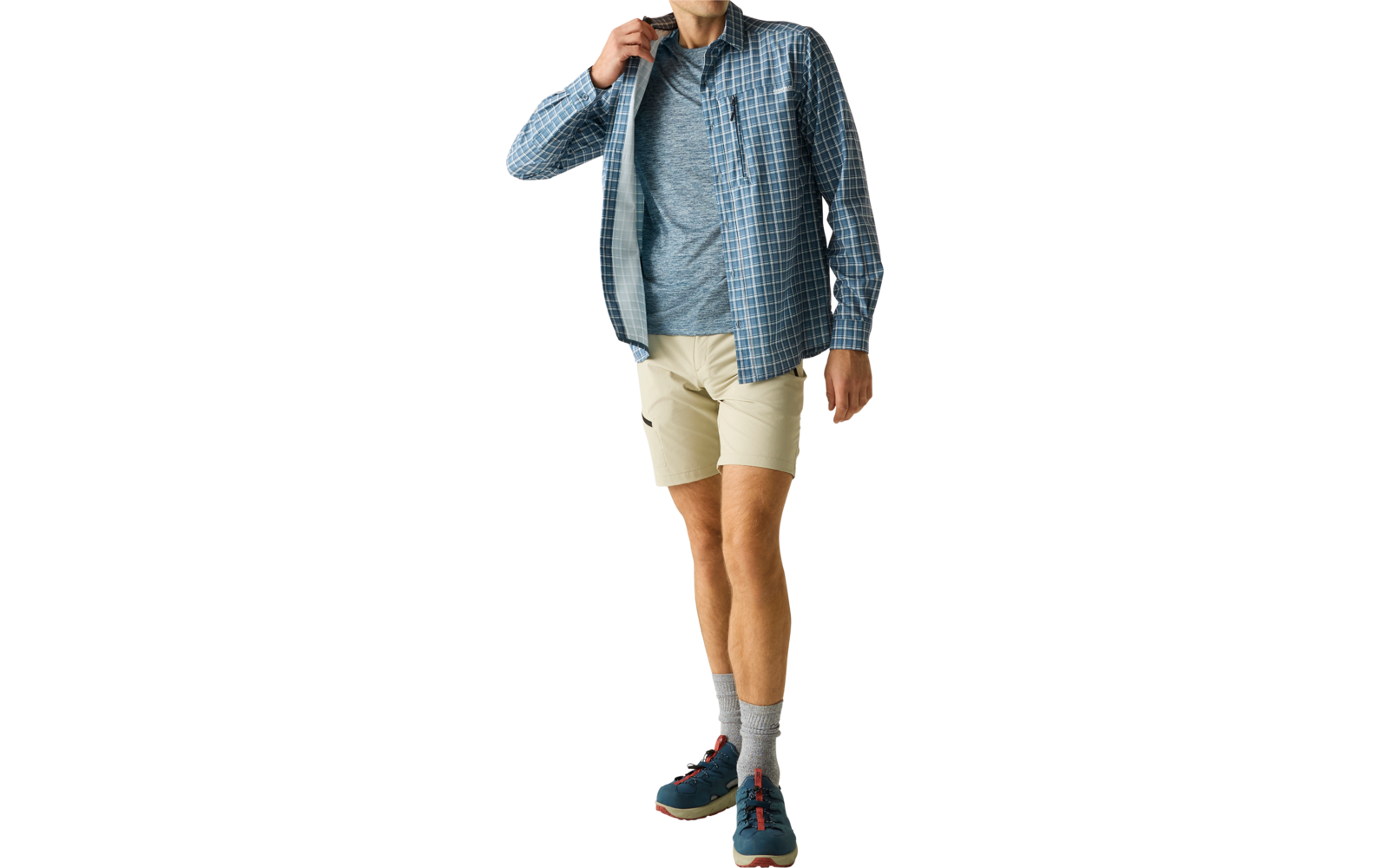 Ein Mann in blau kariertem Hemd über grauem T‑Shirt und beigen Shorts, graue Socken und blaue Sneaker, geht vor hellem Hintergrund und berührt den Hemdkragen