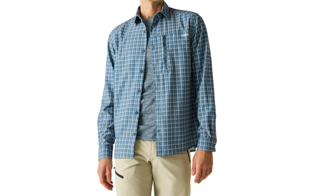 Ein Mann steht vor hellem Hintergrund, trägt ein blau-weiß kariertes Hemd über einem grauen T‑Shirt und helle Hose, blickt nach rechts