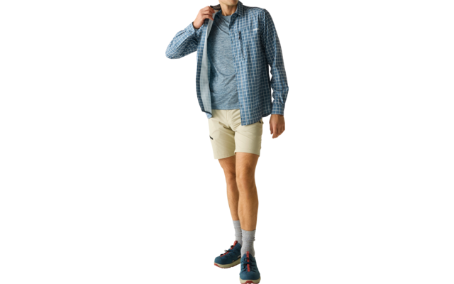 Ein Mann in blau kariertem Hemd über grauem T‑Shirt und beigen Shorts, graue Socken und blaue Sneaker, geht vor hellem Hintergrund und berührt den Hemdkragen