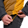 Zwei Hände ziehen an einem gelben Kordelzug am Saum einer zweifarbigen gelben Jacke; schwarze Ärmel und dunkle Hose sichtbar