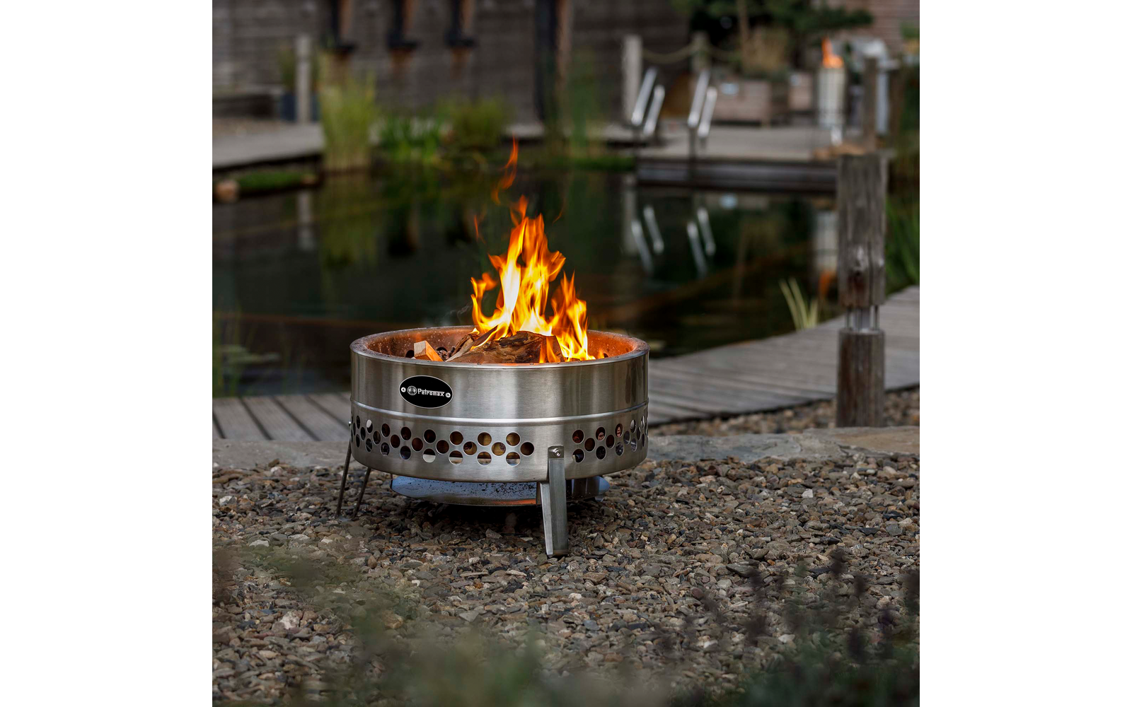 Runde Feuerschale aus Edelstahl mit brennenden Flammen auf Kies vor Holzsteg und Teich