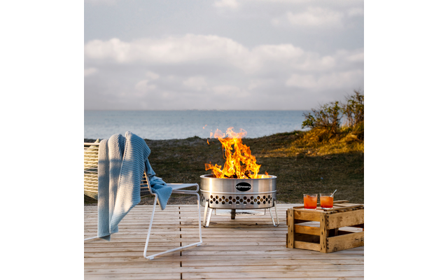 Feuerstelle mit Flammen auf Holzdeck, weißer Stuhl mit blauem Überwurf, Holzkiste mit zwei orangenen Getränken, Meer und bewölkter Himmel im Hintergrund