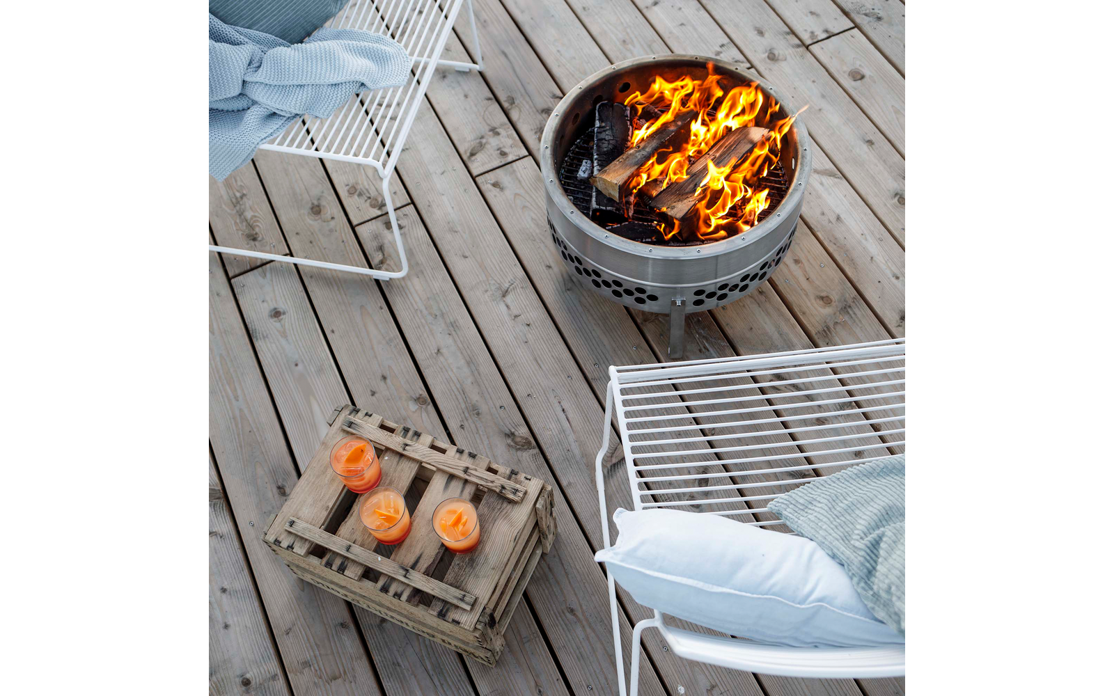 Blick von oben auf ein Holzdeck mit einem brennenden Metallfeuerkorb, zwei weißen Drahtstühlen mit Kissen und Decken und einer Holzkiste mit drei brennenden Kerzen in Glasgefäßen
