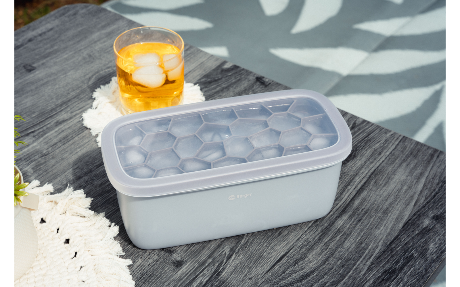 Rechthoekige ijsblokjeshouder met deksel en zichtbare zeskantige vakjes, daarnaast een glas met ijsblokjes en gele drank op een wit gehaakt onderzettertje, donkere houten tafel, zwembadwater en bladschermer in de achtergrond