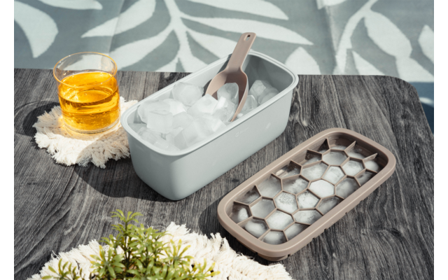 Rechthoekige ijsbak met ijsblokjes en schep, ijsblokjesvorm naast de bak, glas met amberkleurige vloeistof op een onderzetter, tafel met doek en kleine plant