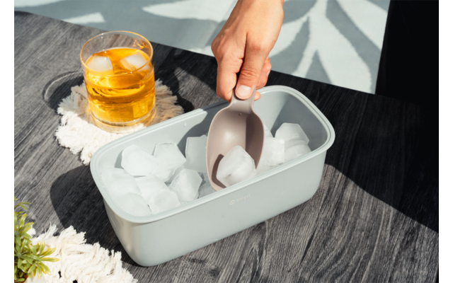 Hand houdt een beige ijslepel boven een lichtgrijze bak met ijsblokjes; daarnaast een glas met een geelachtig drankje en ijs op een witte onderzetter op een donkere houten tafel met schaduwpatroon