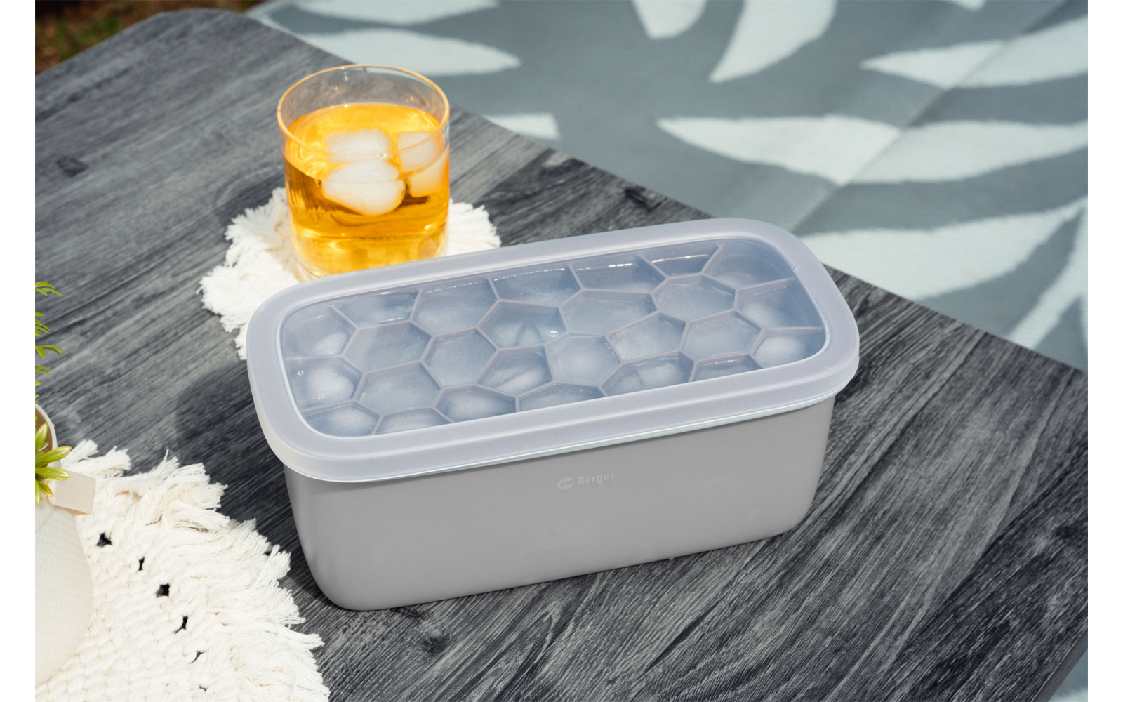 Rechteckiger grauer Behälter mit transparentem Deckel und sechseckigen Fächern auf dunklem Holztisch; daneben ein Glas mit orangefarbener Flüssigkeit und Eiswürfeln, eine weiße gehäkelte Decke und eine Pflanze
