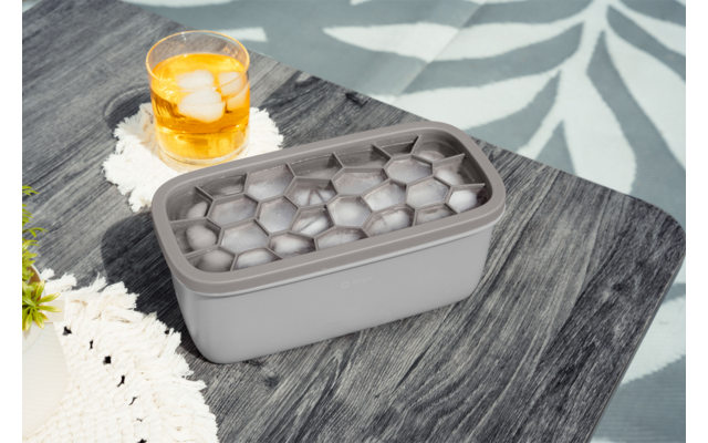 Graues rechteckiges Eiswürfelform mit sechseckigen Eiswürfeln auf einem dunklen Holztisch; daneben ein Glas mit bernsteinfarbener Flüssigkeit und Eis auf einer weißen Häkelunterlage; links ein grünes Pflanzenblatt und ein weißes Häkelstück; im Hinter