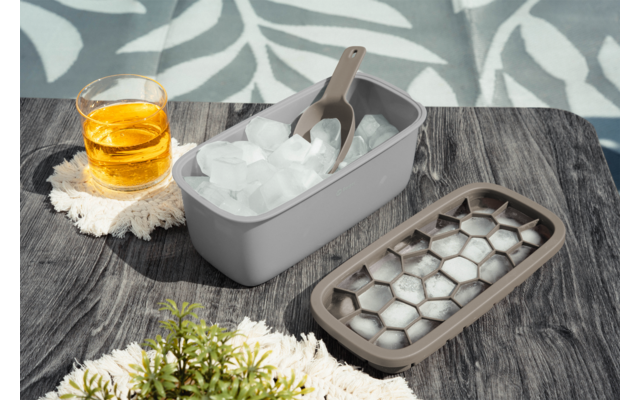 Eiswürfelform mit Eiswürfeln und Schaufel, offener Deckel mit sechseckigen Vertiefungen, Glas mit bernsteinfarbener Flüssigkeit auf einem Tuch, kleine grüne Pflanze, Holztisch und Blatt-Schatten im Hintergrund