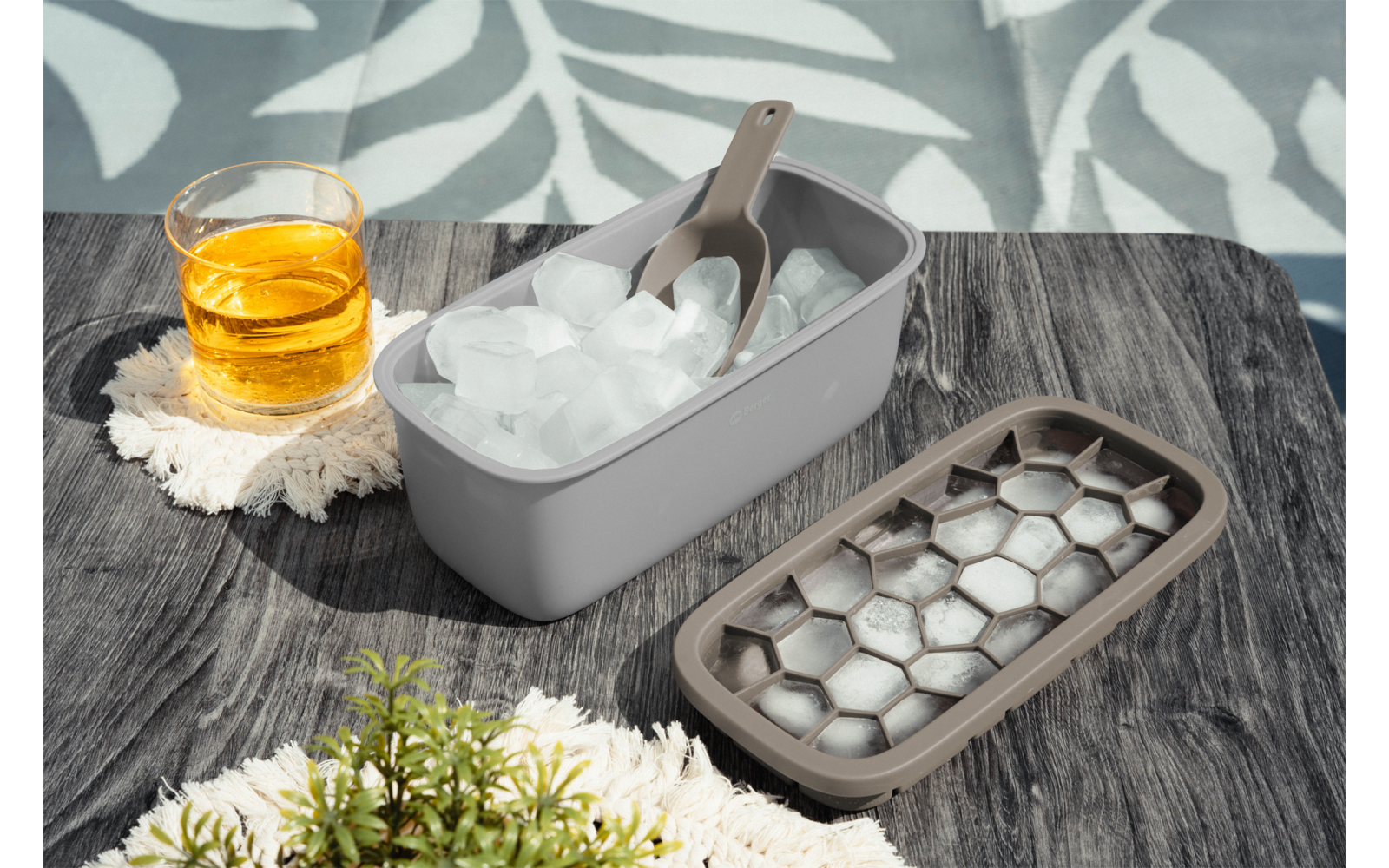 Eiswürfelform mit Eiswürfeln und Schaufel, offener Deckel mit sechseckigen Vertiefungen, Glas mit bernsteinfarbener Flüssigkeit auf einem Tuch, kleine grüne Pflanze, Holztisch und Blatt-Schatten im Hintergrund