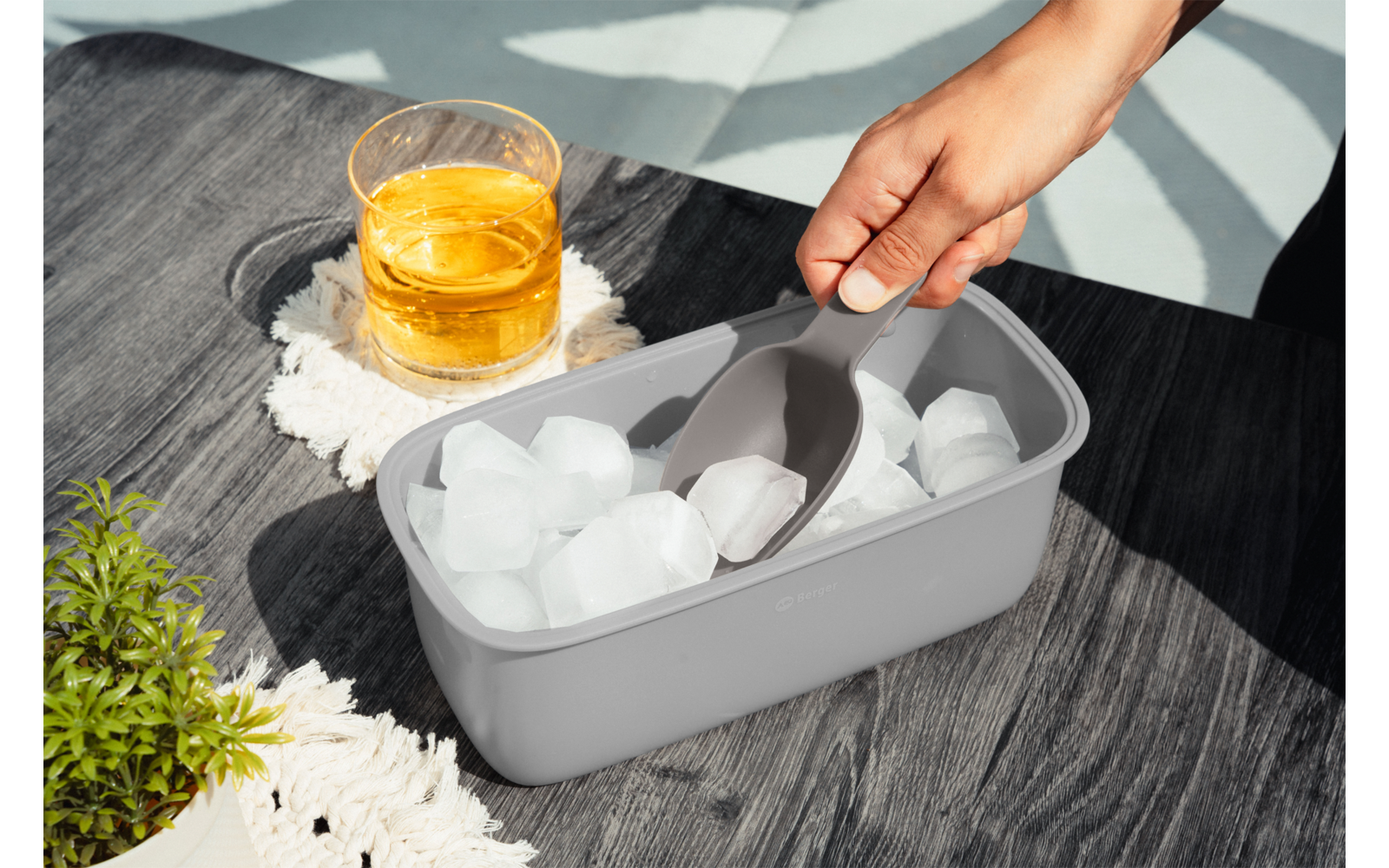 Hand mit Eisschaufel über einem grauen Eisbehälter mit Eiswürfeln auf einem dunklen Holztisch, daneben ein Glas mit bernsteinfarbener Flüssigkeit auf einem Häkeluntersetzer und eine kleine Topfpflanze; Sonnenlicht und Schatten
