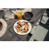 Bord met kleine pannenkoekjes, room, siroop, frambozen en bosbessen; vork op het bord, mes ernaast; glas sinaasappelsap, kom met bosbessen en glazen pot met suiker op een houten tafel; lichtgroene stoelrug zichtbaar