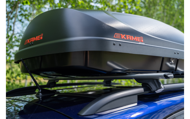 Box da tetto nero con scritta arancione 'EKAME' sul tetto di un'auto blu, alberi sullo sfondo