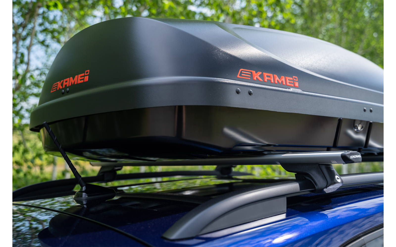 Box da tetto nero con scritta arancione 'EKAME' sul tetto di un'auto blu, alberi sullo sfondo