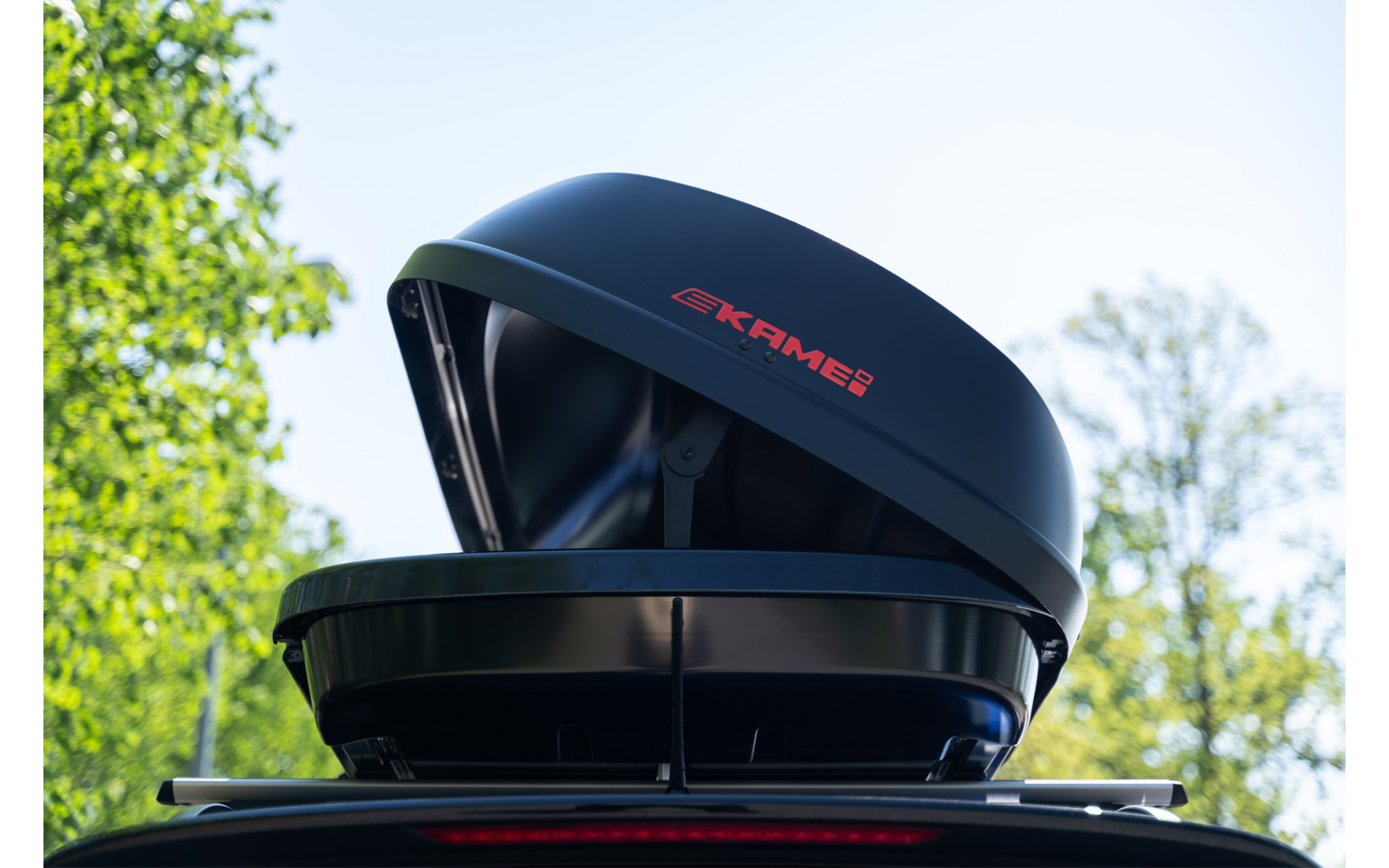 Baule da tetto nero aperto con logo rosso KAMEI montato sul tetto di un'auto, alberi e cielo sullo sfondo