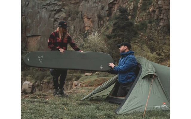 Zwei Personen sitzen neben einem Zelt und halten zusammen ein großes Surfbrett vor einer Felswand