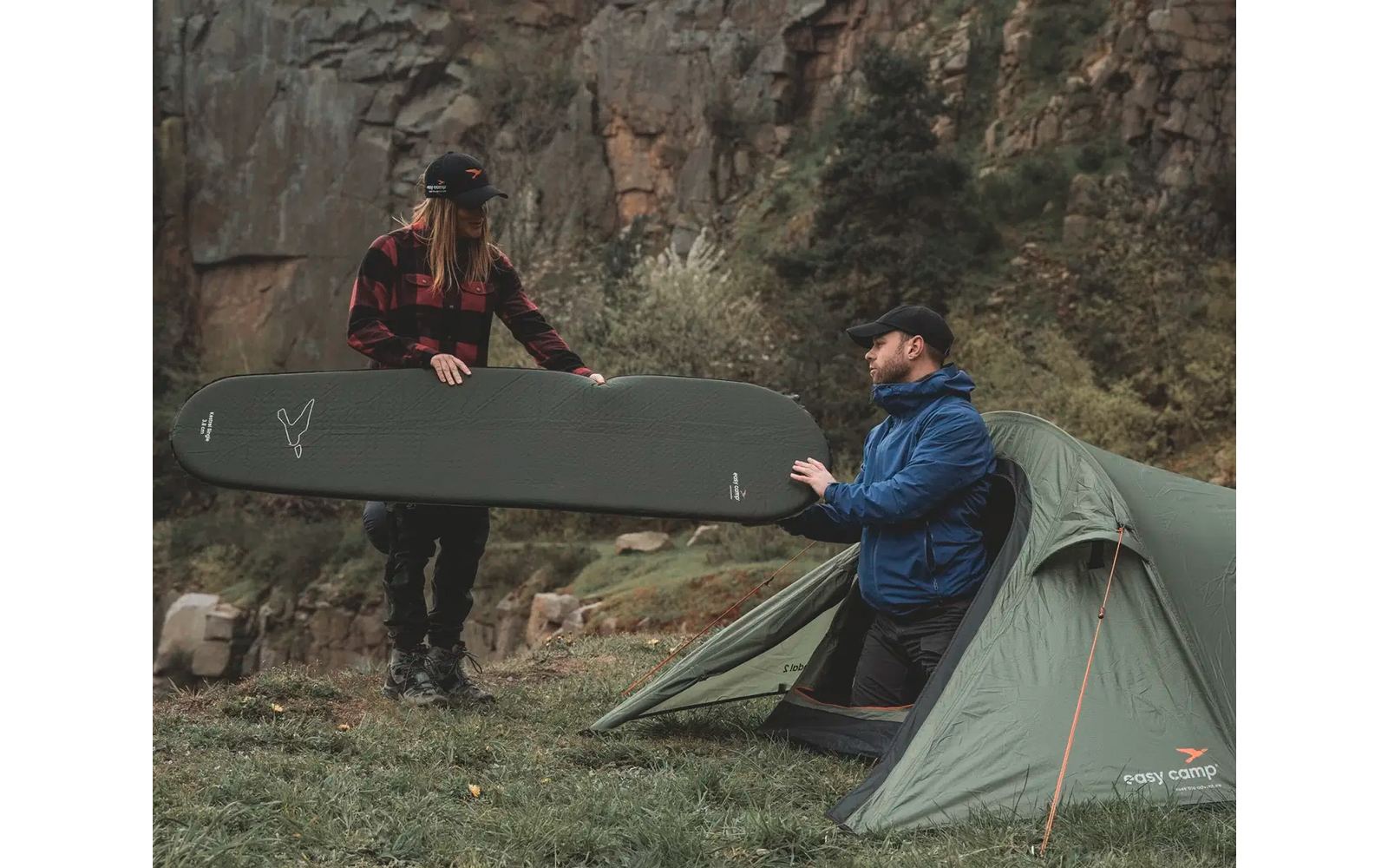 Zwei Personen sitzen neben einem Zelt und halten zusammen ein großes Surfbrett vor einer Felswand