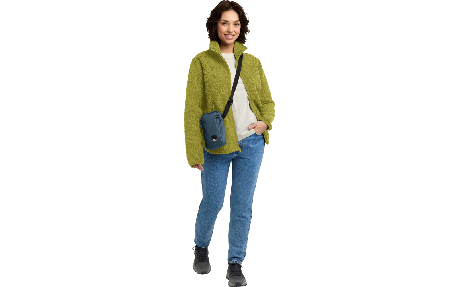 Persoon staat, draagt groene fleecejas, wit sweatshirt, blauwe spijkerbroek, zwarte schoenen en een blauwe schoudertas schuin over de borst; glimlachend
