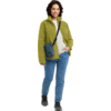 Persoon staat, draagt groene fleecejas, wit sweatshirt, blauwe spijkerbroek, zwarte schoenen en een blauwe schoudertas schuin over de borst; glimlachend
