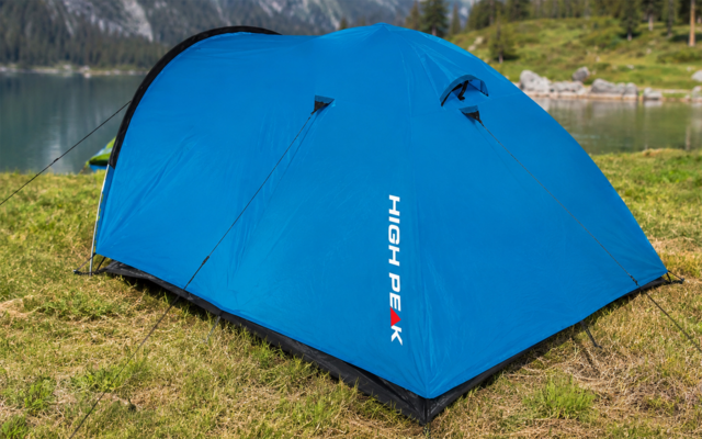 Blaues Kuppelzelt mit weißem "HIGH PEAK"-Schriftzug, abgespannt auf Gras nahe einem See, im Hintergrund Berge und Bäume