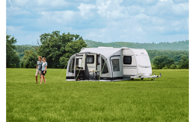 Wohnwagen mit angebautem Vorzelt auf einer grünen Wiese, zwei Personen gehen links davon, Baumreihe und bewölkter Himmel im Hintergrund