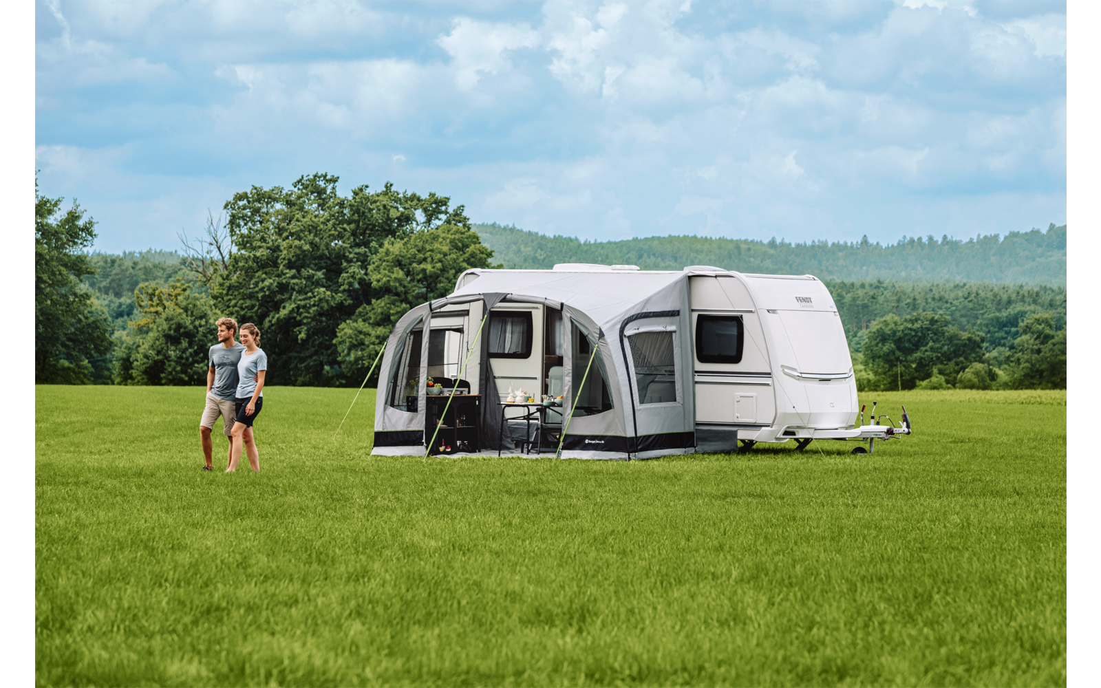 Wohnwagen mit angebautem Vorzelt auf einer grünen Wiese, zwei Personen gehen links davon, Baumreihe und bewölkter Himmel im Hintergrund