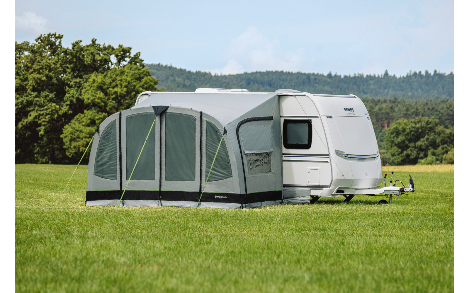 Weißer Wohnwagen mit grauem Vorzelt auf grüner Wiese, im Hintergrund Bäume und bewaldete Hügel unter hellem Himmel