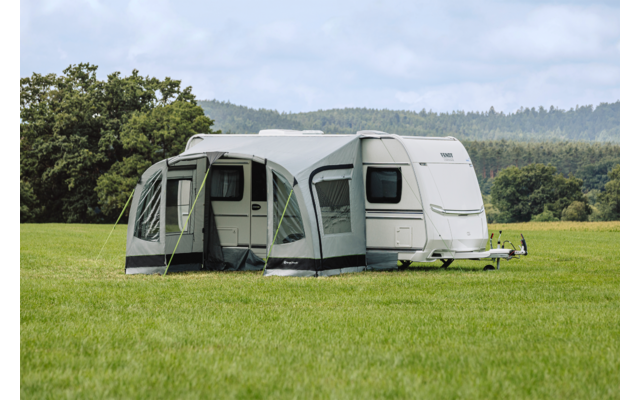 Weißer Wohnwagen mit grauem Vorzelt auf einer großen Wiese, im Hintergrund Bäume und bewaldete Hügel unter bedecktem Himmel