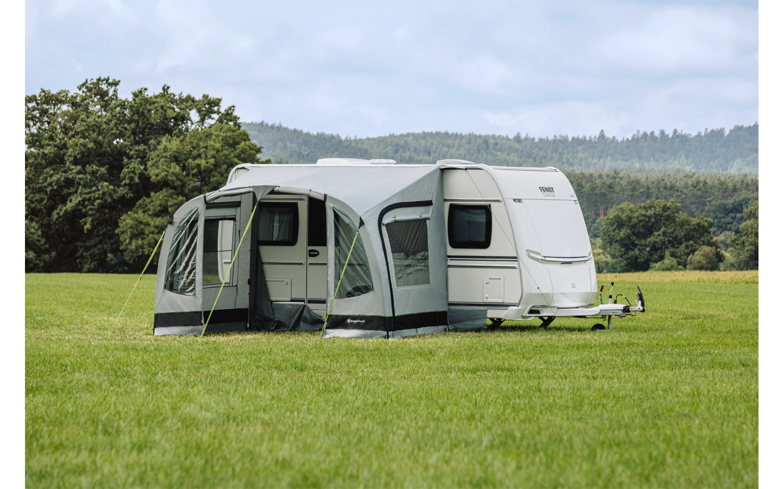 Weißer Wohnwagen mit grauem Vorzelt auf einer großen Wiese, im Hintergrund Bäume und bewaldete Hügel unter bedecktem Himmel