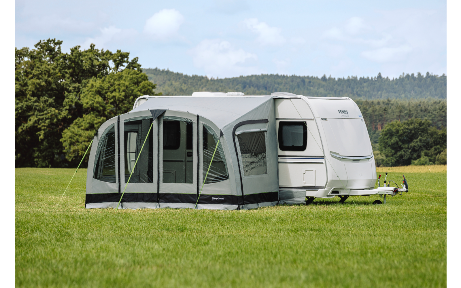Weißer Wohnwagen mit grauem Vorzelt auf einer grünen Wiese, Bäume und Hügel im Hintergrund, blauer Himmel mit Wolken