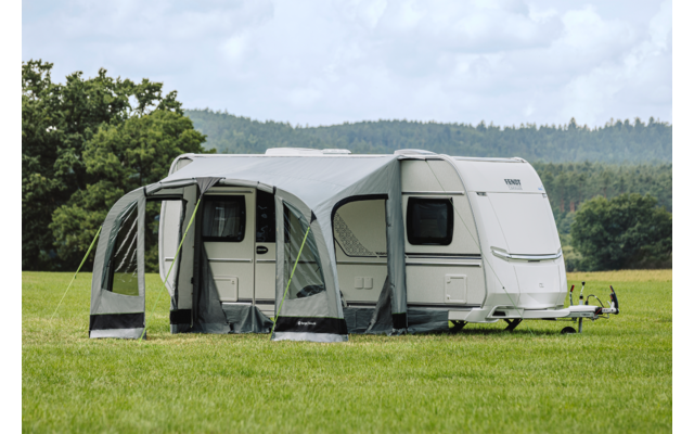 Weißer Wohnwagen mit angebautem Vorzelt auf einer Wiese, im Hintergrund Bäume und bewölkter Himmel