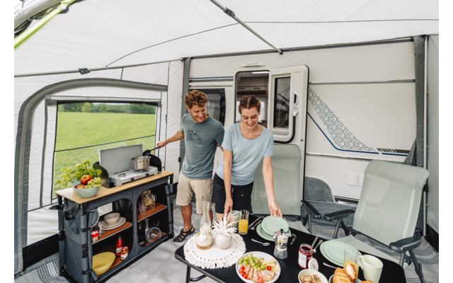 Zwei Personen im Vorzelt eines Wohnmobils neben einem Tisch mit gedecktem Frühstück, Camping-Küchenschrank und Klappstühlen; draußen Wiese sichtbar