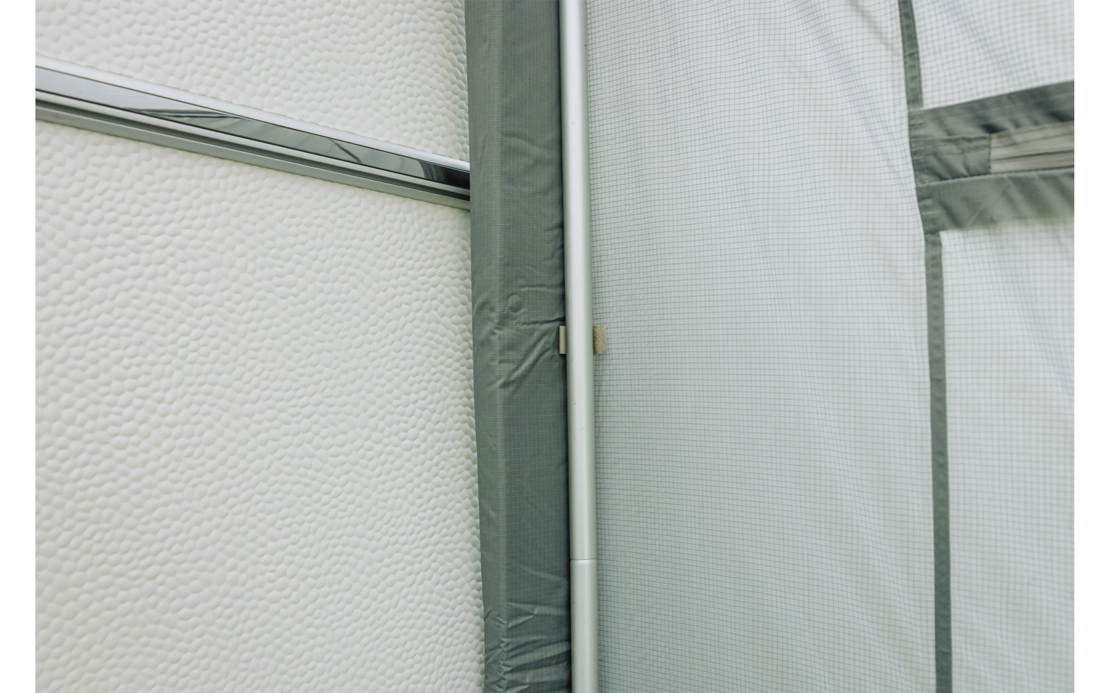 Texturierte weiße Fläche links, vertikale weiße Metallstange nahe der Bildmitte, graugrüne Stoffbespannung mit Nähten und dunklerem Rand rechts