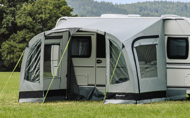 Weißer Wohnwagen mit grauem Vorzelt auf einer großen Wiese, im Hintergrund Bäume und bewaldete Hügel unter bedecktem Himmel