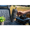 Hand holding a dark gray travel tumbler with handle and 'Camp Life' print over a black folding table; potted plant left, portable gas stove right, blurred grass background