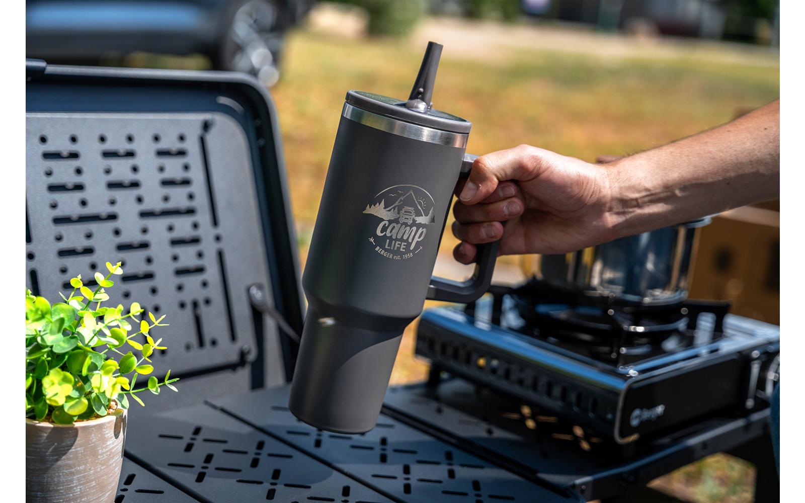 Hand holding a dark gray travel tumbler with handle and 'Camp Life' print over a black folding table; potted plant left, portable gas stove right, blurred grass background