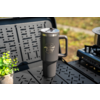 Gray insulated travel mug with handle and "Camp Life" logo on a black perforated camping table; small potted green plant to the left, camping stove to the right