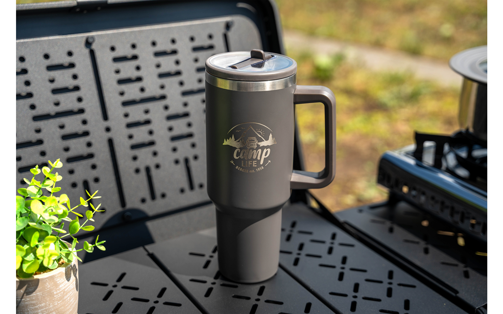Gray insulated travel mug with handle and "Camp Life" logo on a black perforated camping table; small potted green plant to the left, camping stove to the right
