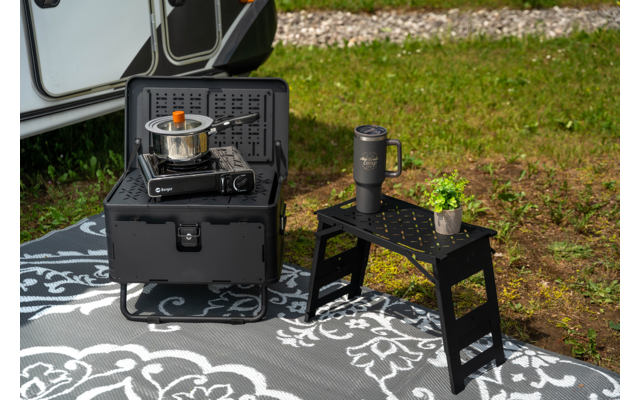 Gas stove in a black folding case with a small pot, next to a black two-step stool with a travel mug and a small potted plant on a patterned outdoor mat beside a caravan