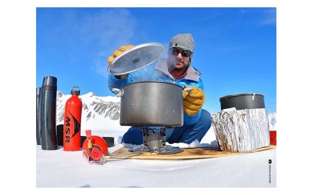 Person mit Mütze, Sonnenbrille und Handschuhen hebt einen Topfdeckel über einem tragbaren Kocher auf Schnee; im Hintergrund schneebedeckte Berge, links Thermoskanne und rote MSR‑Flasche, rechts weiterer Topf auf Aluminiumfolie
