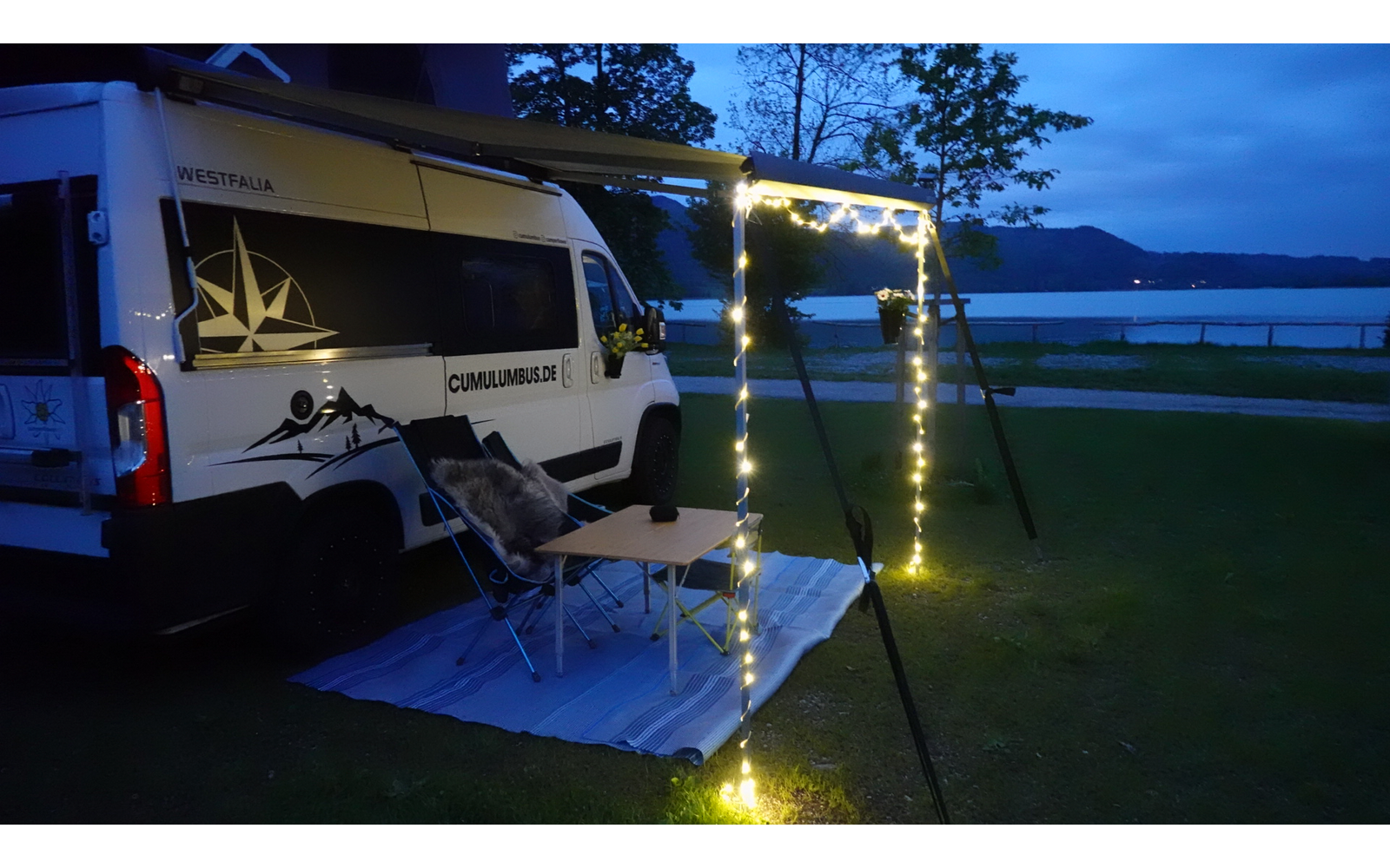 Wohnmobil mit geöffnetem Seitenfenster und Markise an einem Seeufer bei Abenddämmerung, mit Lichterkette und Hängematte im Gras