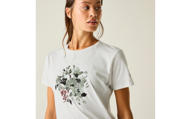 Person von der Brust aufwärts, trägt weißes T‑Shirt mit grüner und schwarzer Blumenillustration und roten Akzenten, Haare zurückgebunden, blickt nach rechts, heller einfarbiger Hintergrund