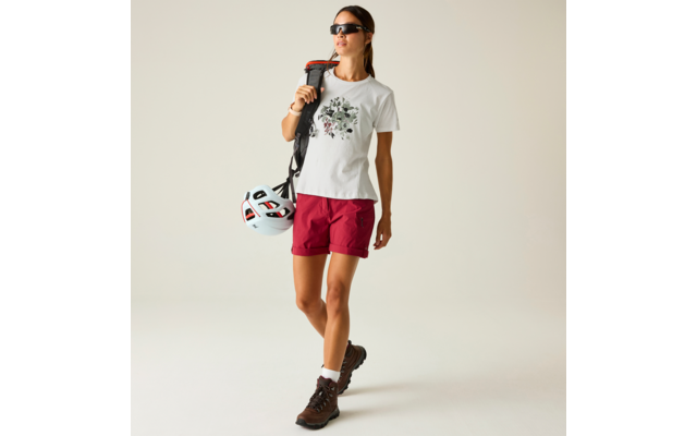 Person mit Sonnenbrille, weißem T-Shirt mit Blumenmotiv, roten Shorts, Wanderschuhen, trägt Rucksack über einer Schulter und hält einen Fahrradhelm vor hellem Hintergrund