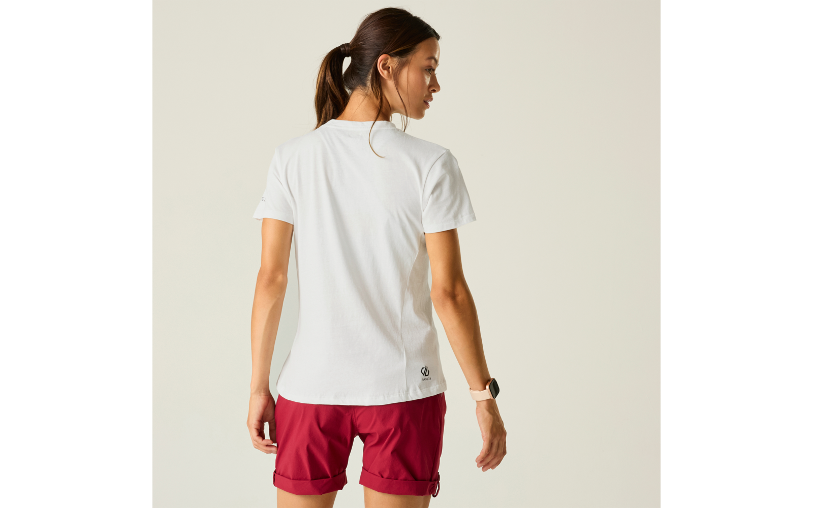 Person mit dem Rücken zur Kamera, trägt weißes T‑Shirt und rote Shorts, Haare zu Pferdeschwanz, heller einfarbiger Hintergrund