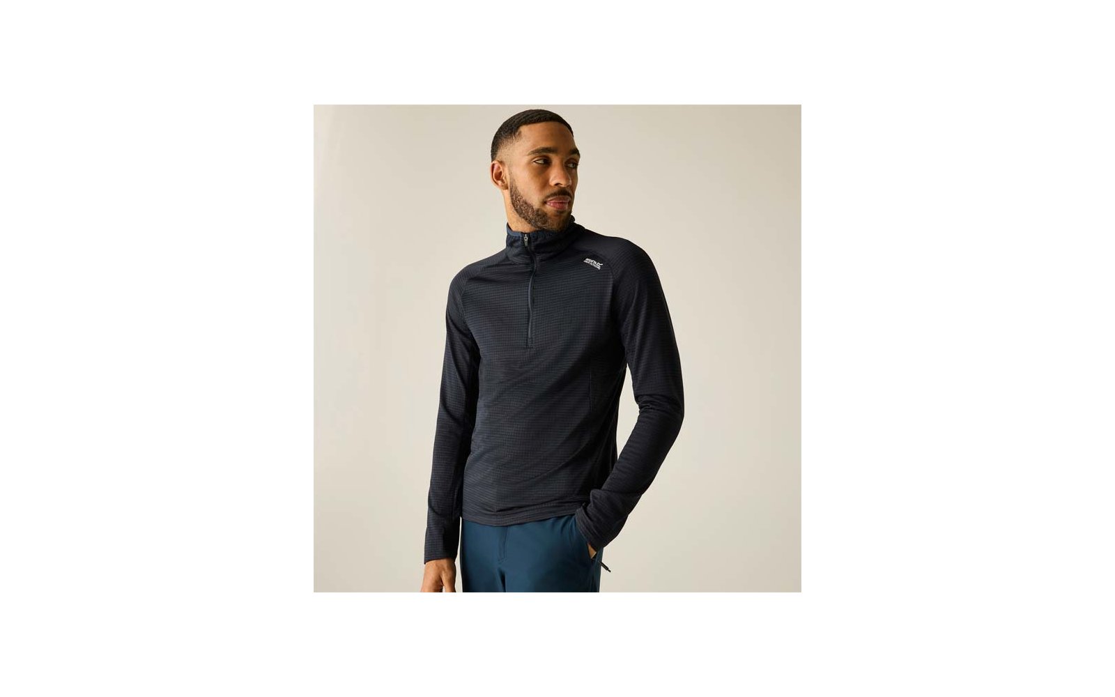 Personne portant un haut noir à manches longues avec une fermeture éclair courte et petit logo blanc sur la poitrine, pantalon bleu, debout sur fond clair et regardant vers la droite