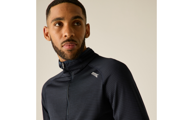 Homme aux cheveux courts et barbe, porte un haut zippé sombre à texture fine avec un petit logo blanc sur la poitrine, regarde sur le côté, fond uni clair