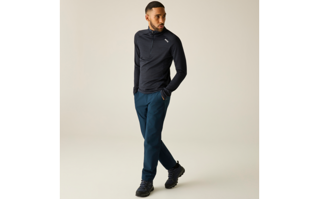 Homme portant un haut sombre à demi-fermeture éclair à manches longues avec petit logo sur la poitrine, pantalon bleu et chaussures de randonnée noires, mains dans les poches, debout jambes croisées sur fond clair