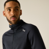 Homme aux cheveux courts et barbe, porte un haut zippé sombre à texture fine avec un petit logo blanc sur la poitrine, regarde sur le côté, fond uni clair