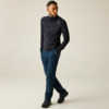 Homme portant un haut sombre à demi-fermeture éclair à manches longues avec petit logo sur la poitrine, pantalon bleu et chaussures de randonnée noires, mains dans les poches, debout jambes croisées sur fond clair